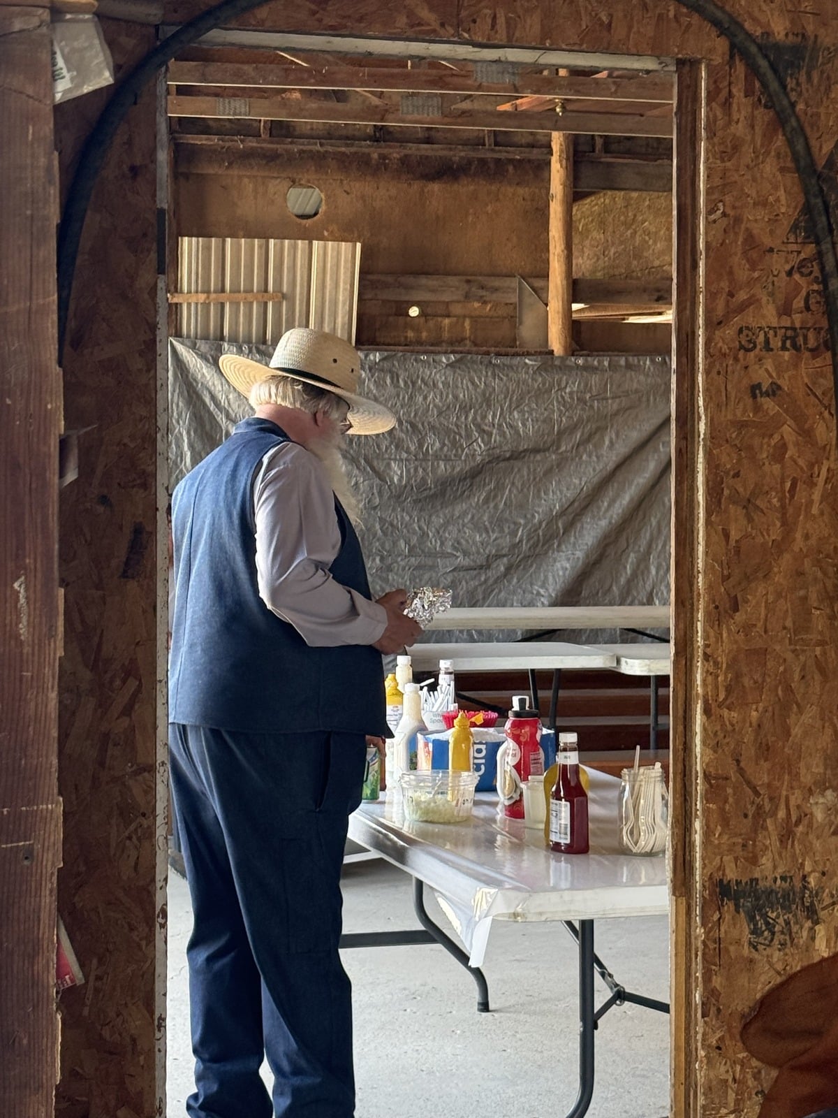 Older Amish man in straw hat standing at condiment table inside barn framed by rough doorway opening