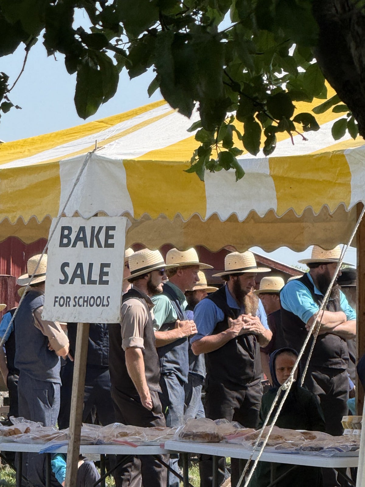 Bake sale for schools sign in front of Amish men in straw hats under yellow and white striped tent