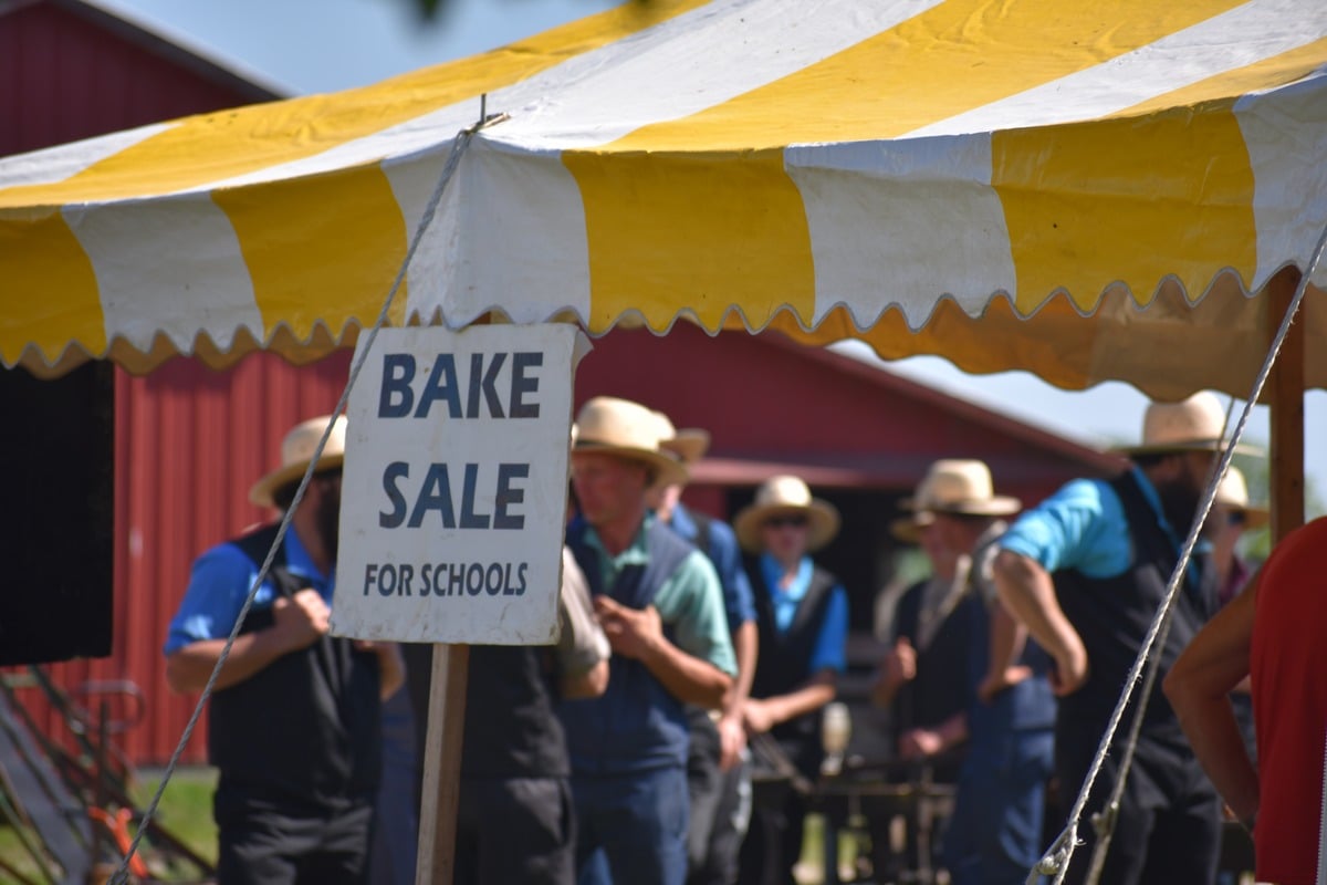 Bake sale for schools sign on post under yellow and white striped tent with group of Amish men in straw hats and red barn behind