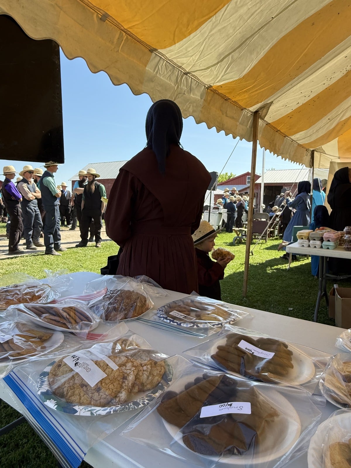 Baked goods in bags on table under auction tent with Amish woman and child watching crowd behind