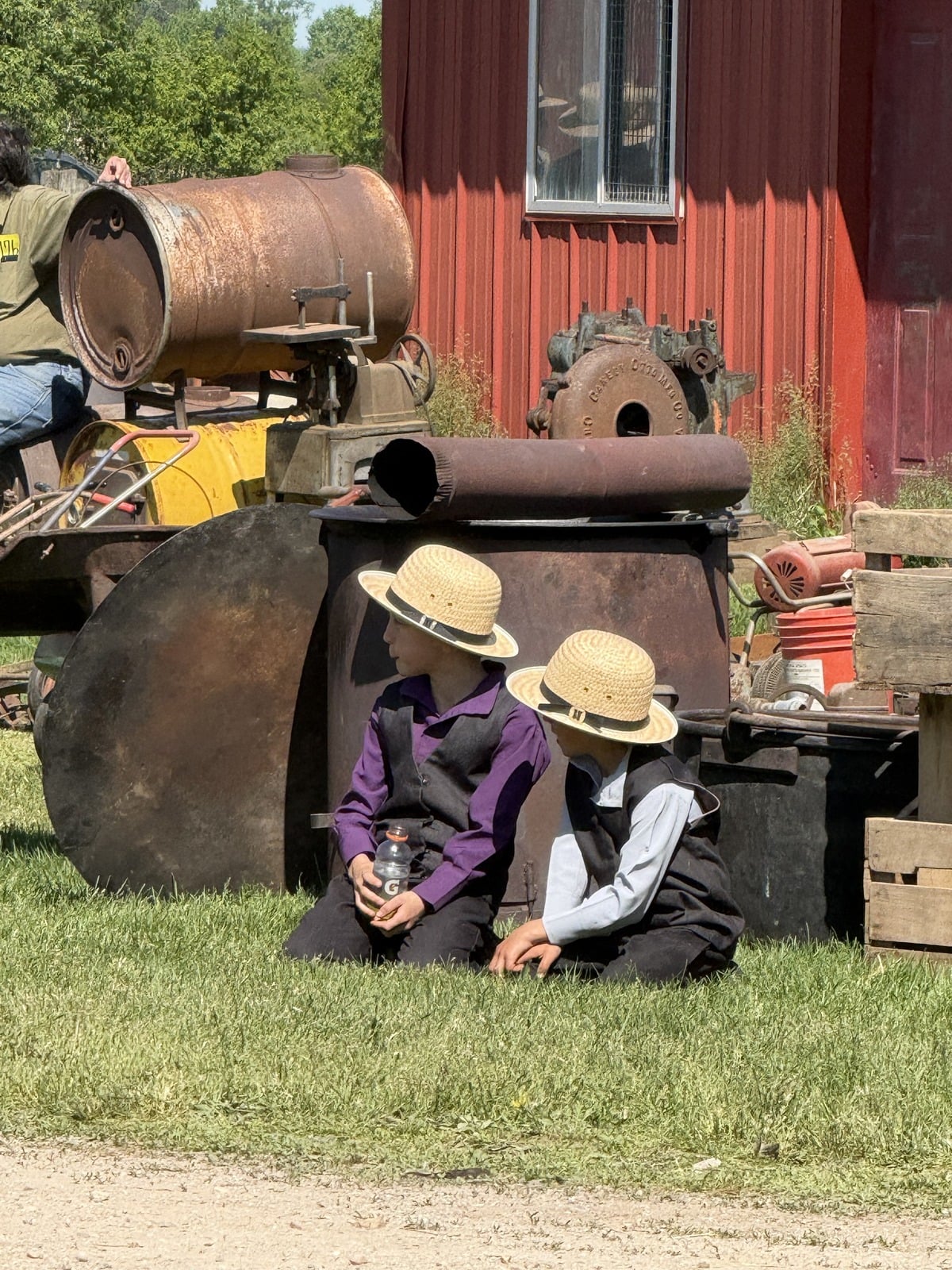 Two young Amish boys in straw hats sitting in grass beside rusty farm machinery at red barn