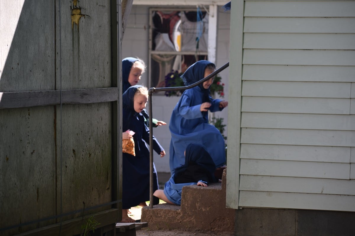 Three young Amish girls in dark dresses running through gap between buildings one clutching a bag of chips