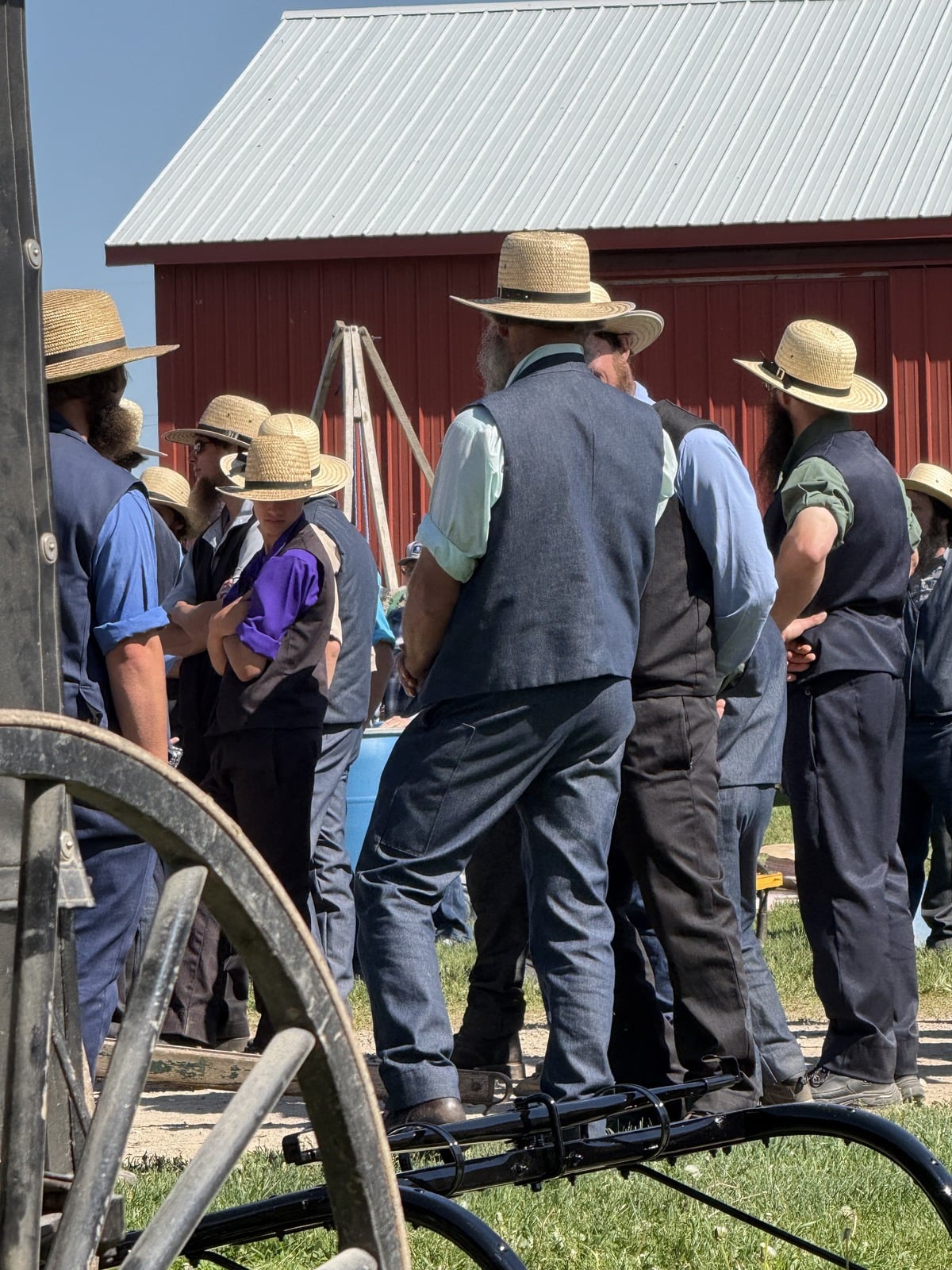 Group of Amish men in straw hats gathered at auction with buggy wheel in foreground and red barn behind