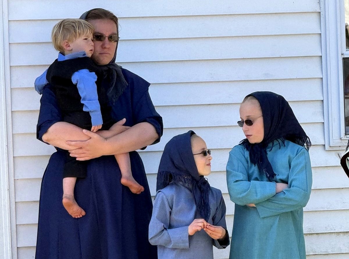 Woman in a navy dress and headscarf holds a young blond boy, while two girls in matching headscarves and modest dresses stand beside them against a white siding wall.