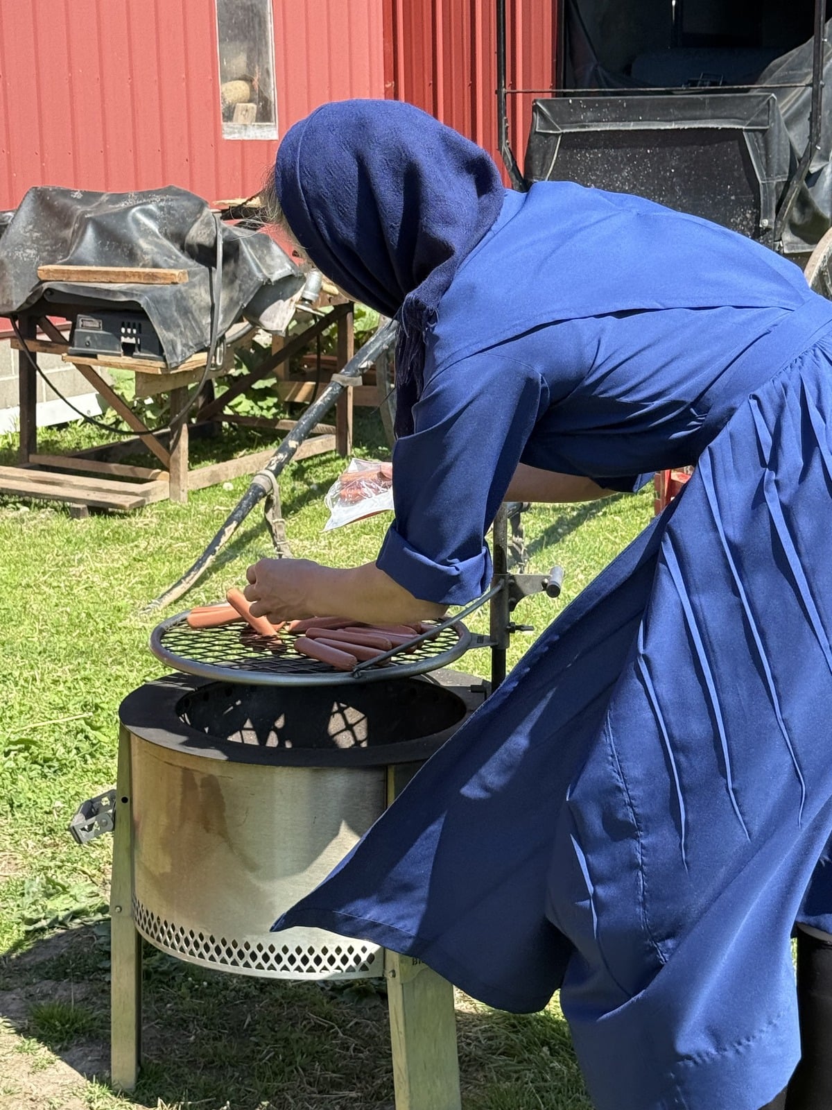 Amish woman in blue dress leaning over outdoor grill tending hot dogs with red barn and buggies behind