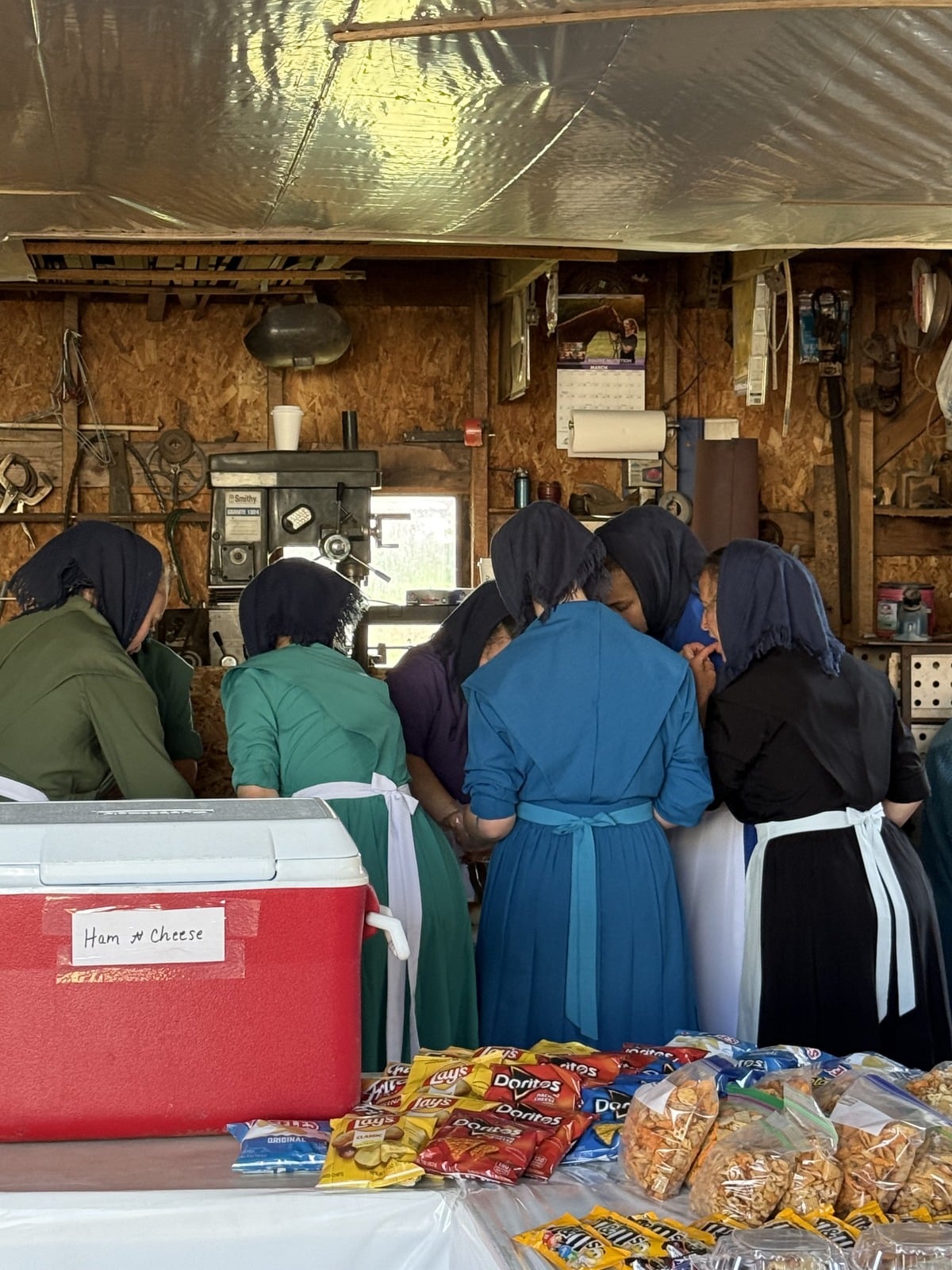 Group of Amish women and girls at food stand inside barn with Lays Doritos chips and ham and cheese cooler