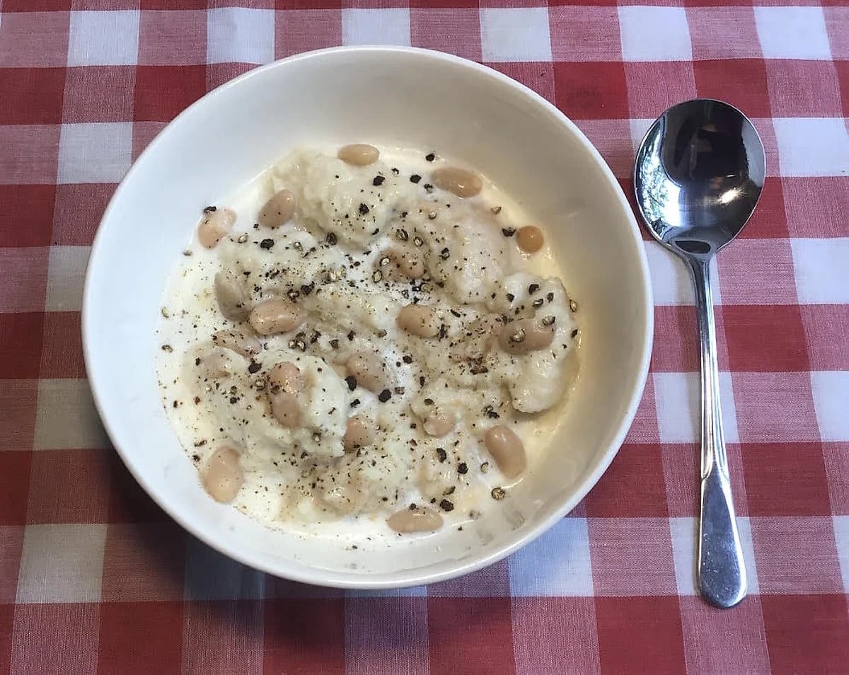 White bowl of Amish bread soup with navy beans and black pepper on a red checkered cloth, spoon alongside