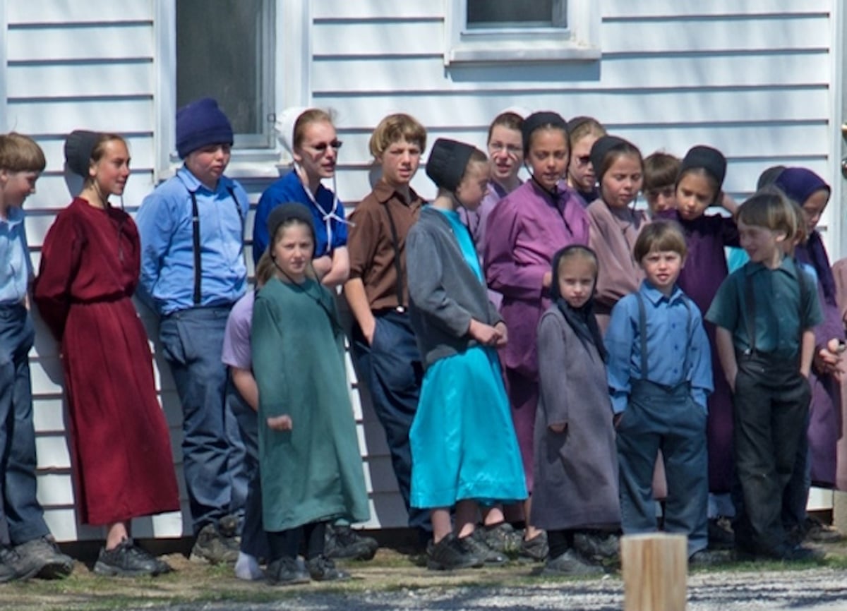 Amish school children gathered outside a schoolhouse A large group of Amish school children in colorful dresses and shirts stand together against the side of a white schoolhouse