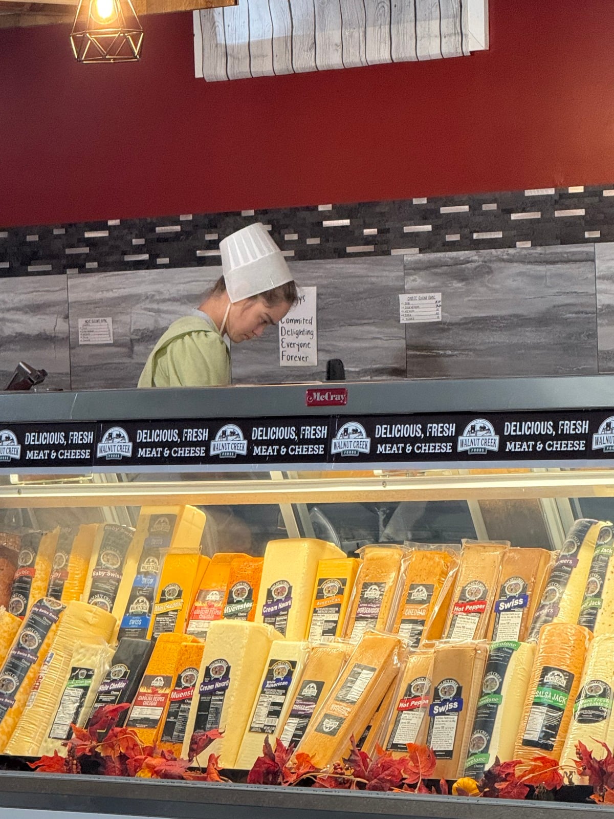 Amish woman in a white cap working behind a deli counter above a case of cheeses at The Farmers' Creamery store in Mio, Michigan