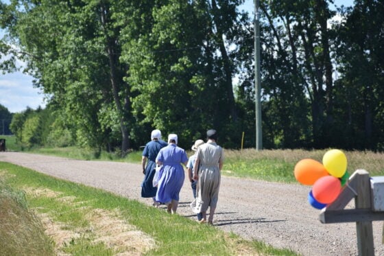 Group of people in long, pastel dresses walking down a gravel rural road, with balloons tied to a post on the right.