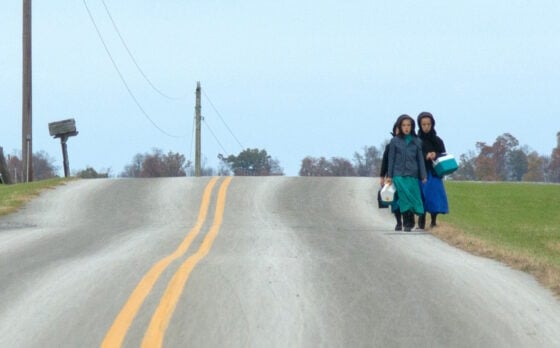 Three people walk up a gently rising country road, carrying plastic coolers and jugs on a rural day.