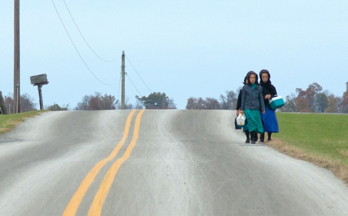 Two Amish girls walk along a rural road carrying lunch coolers on an overcast fall day