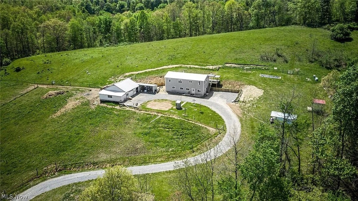 Aerial view of a rural property with curved driveway, two white buildings, and green pastures bordered by trees in the background.
