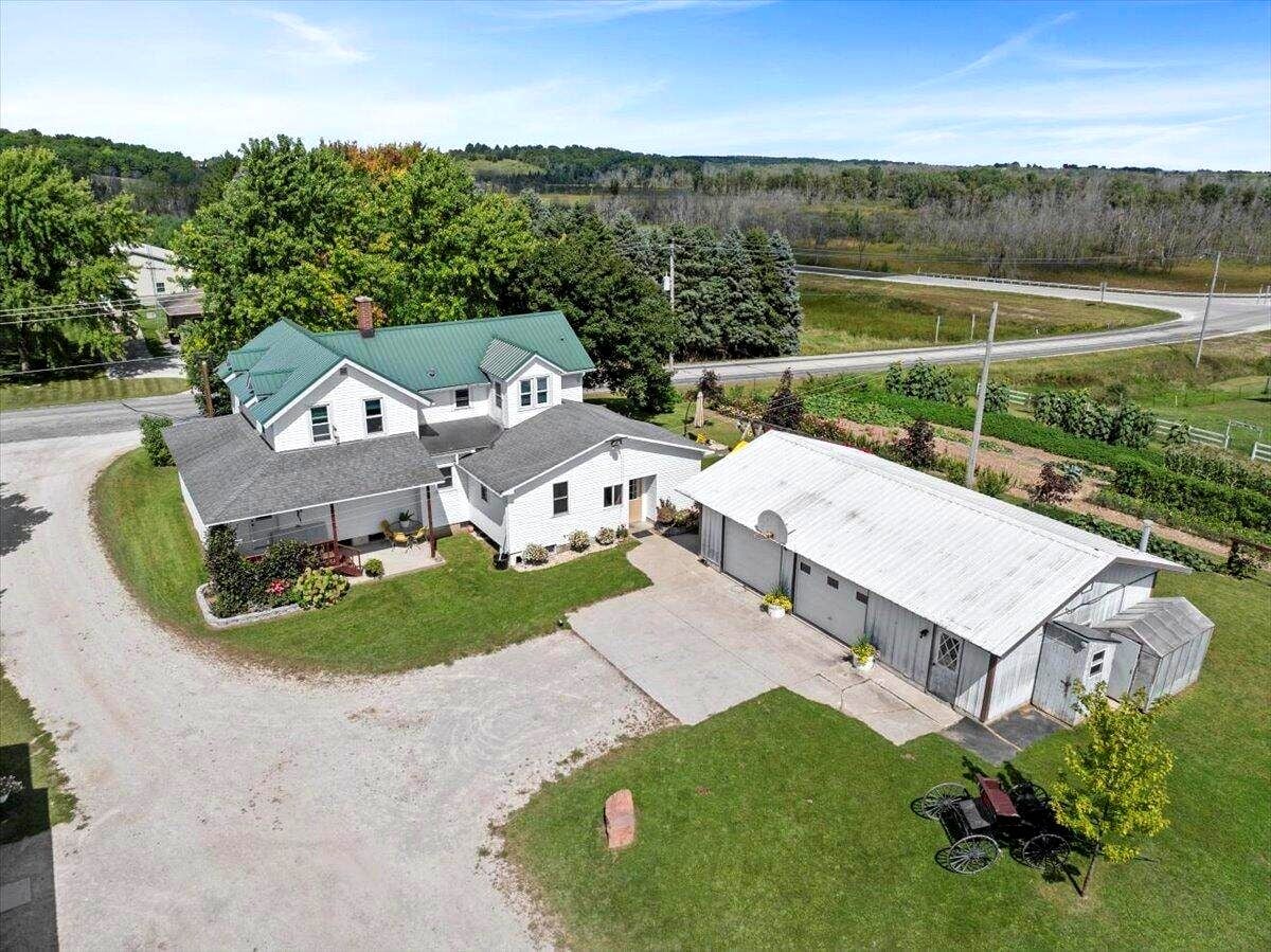 Aerial view of a white two-story house with a green metal roof, attached porch, and a large white barn on a gravel driveway in a rural setting.