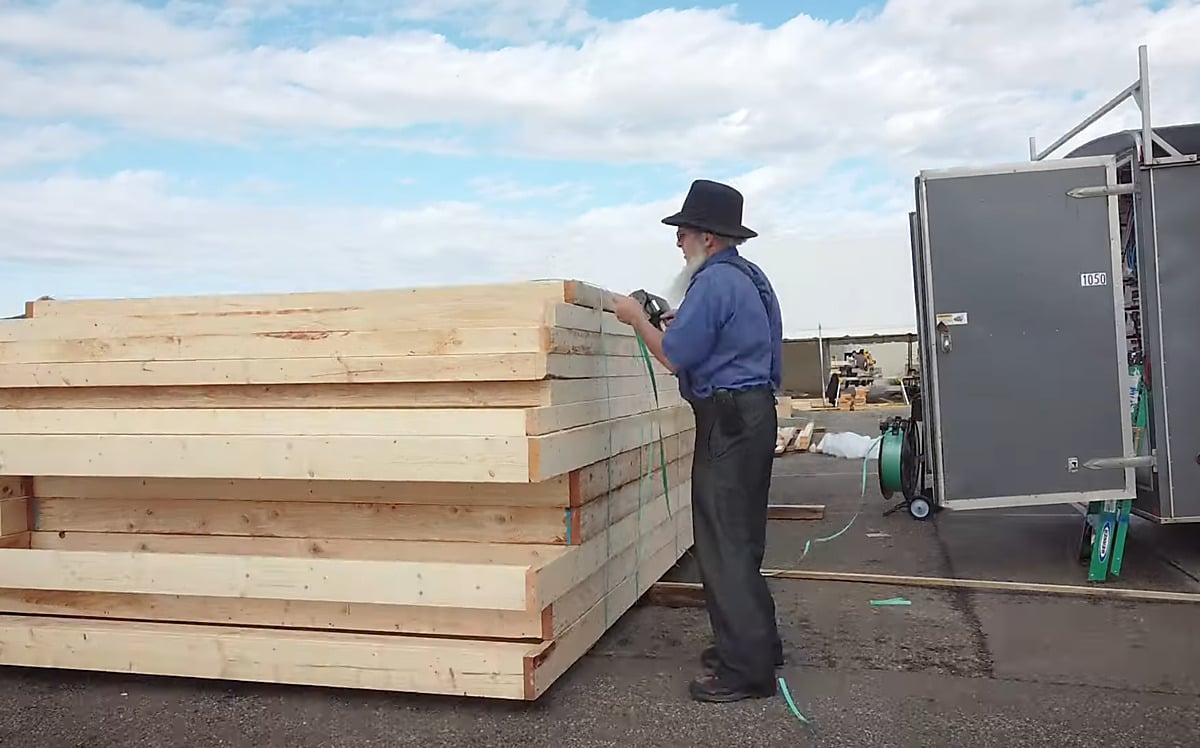Amish builder banding wall panels in Charm, Ohio Amish man in a black hat and blue shirt banding a stack of framed wall panels beside a gray work trailer