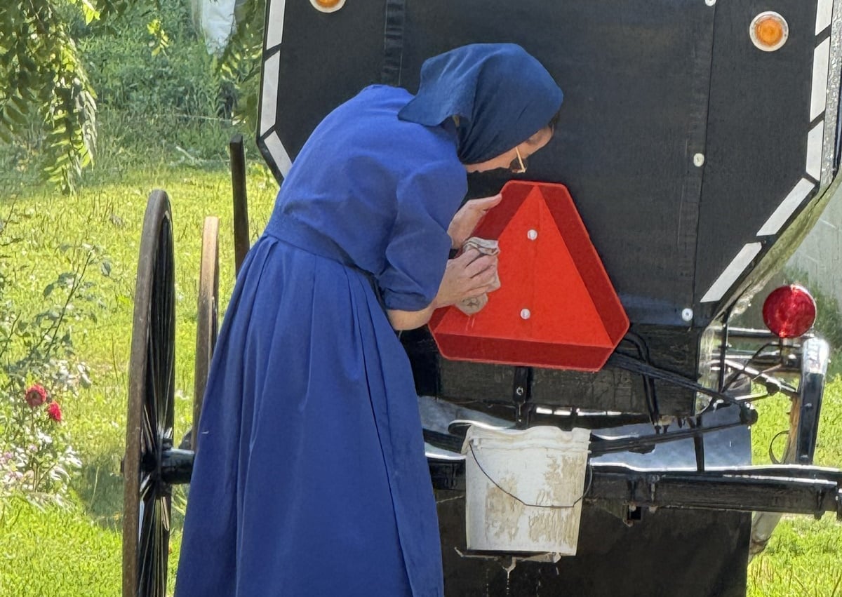 An Amish woman in a blue dress and headscarf wipes down the orange slow-moving vehicle triangle on the back of a buggy, with a soapy bucket hanging from the rear step