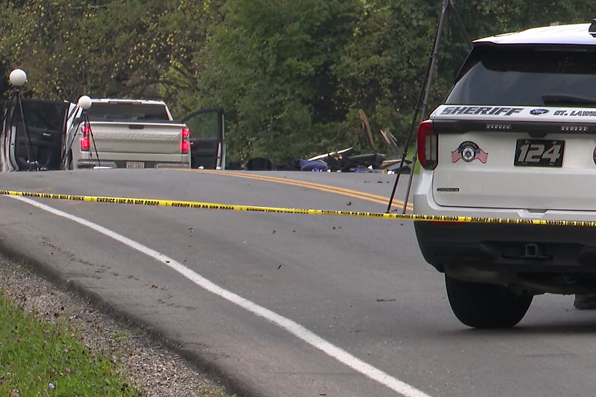 Sheriff's tape across a rural road with crash debris and a pickup truck in the background