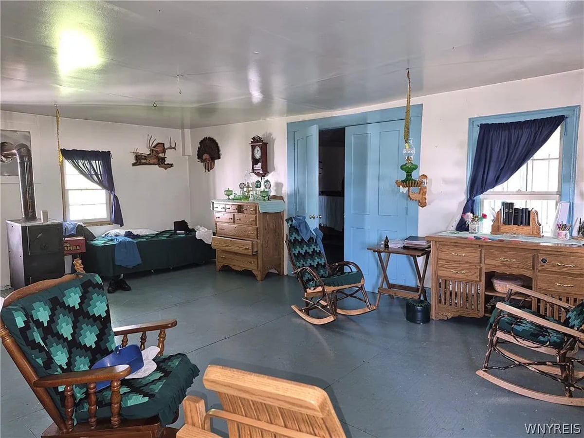 Wide view of the Amish home's front room with a bed, wood stove, dresser, and blue doors
