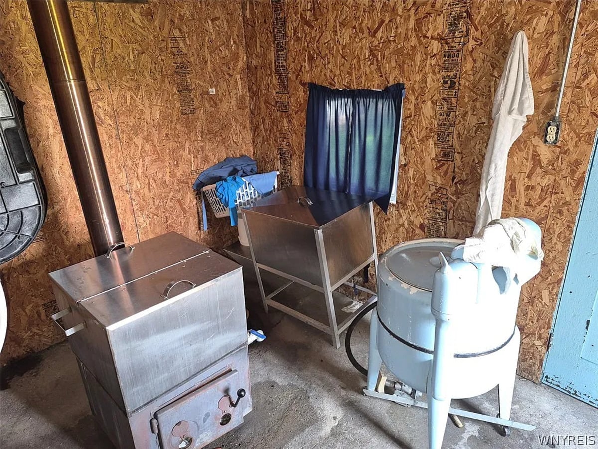 Wringer washer on legs beside two metal water-heating tanks and a small wood stove in an Amish wash house
