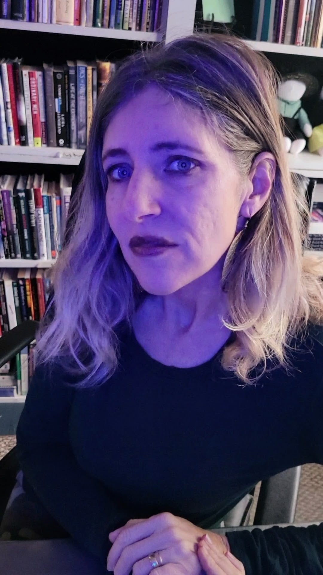 Portrait of a woman with shoulder-length blond hair wearing a black top, seated at a desk with bookshelves in the background under purple lighting.