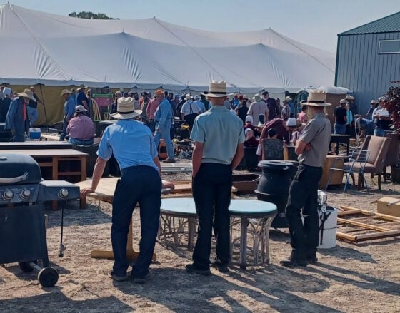 People in hats browse a busy outdoor yard sale under large white tents, with tables, chairs, and scattered furniture and appliances.