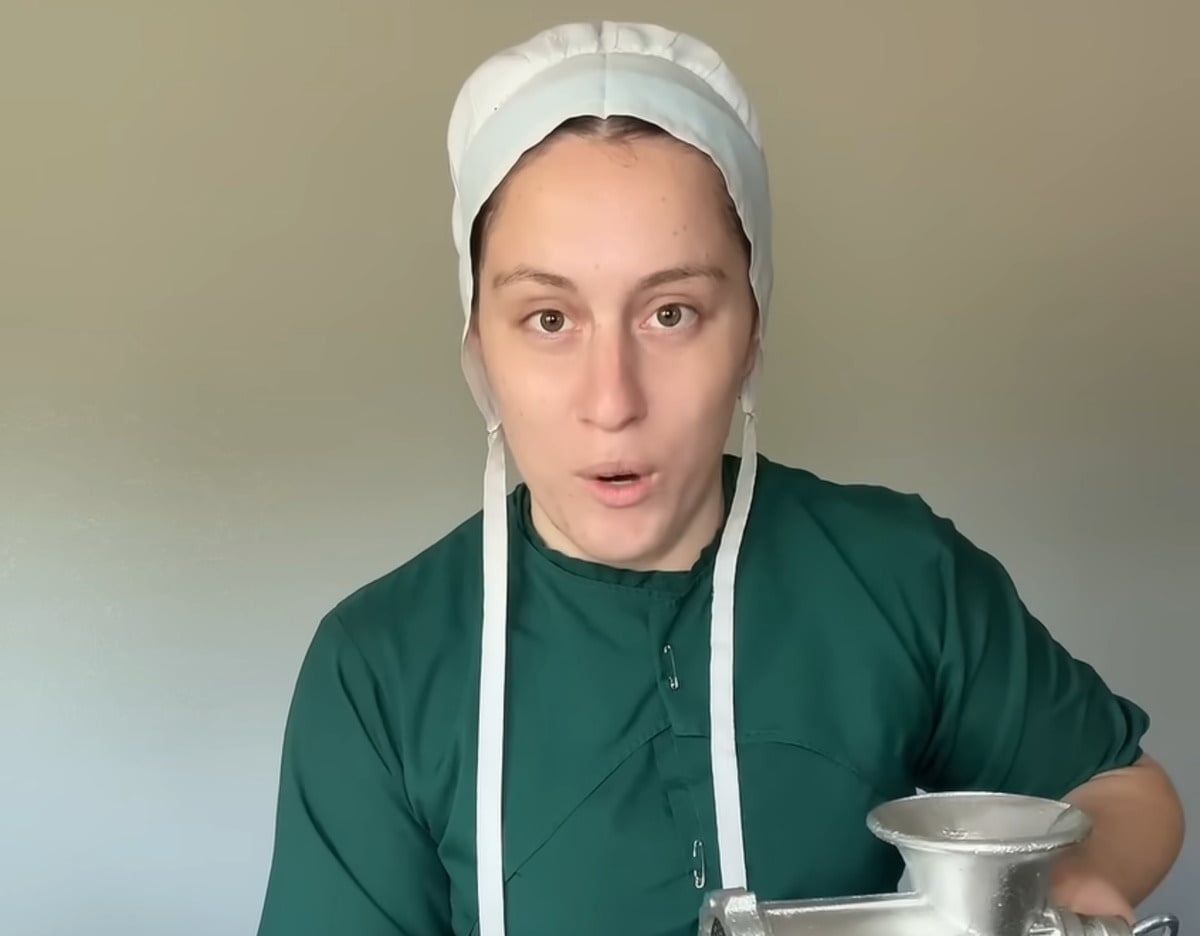 Lovina Zook in a green Amish dress and white covering, looking directly at the camera while holding a hand-crank meat grinder