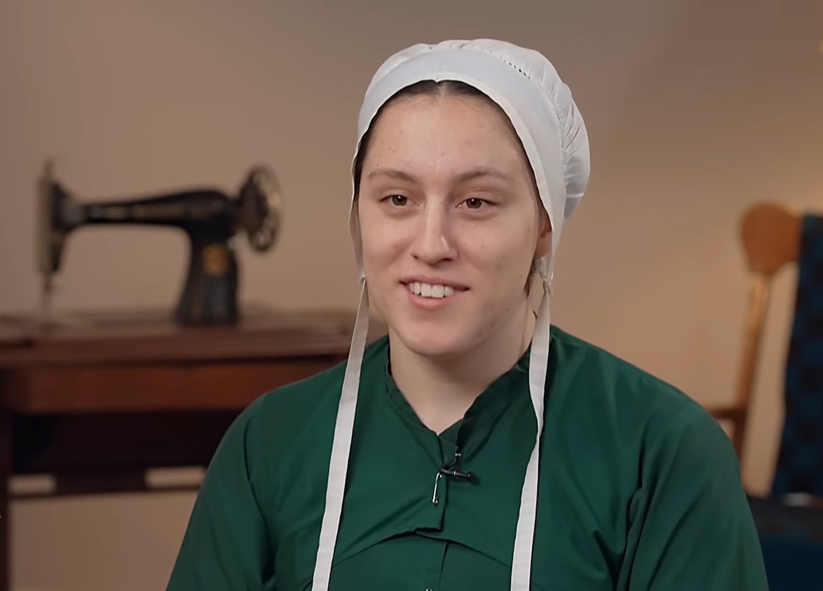 Lovina Zook in a green dress and white covering, seated in front of a treadle sewing machine during her CBS Sunday Morning interview