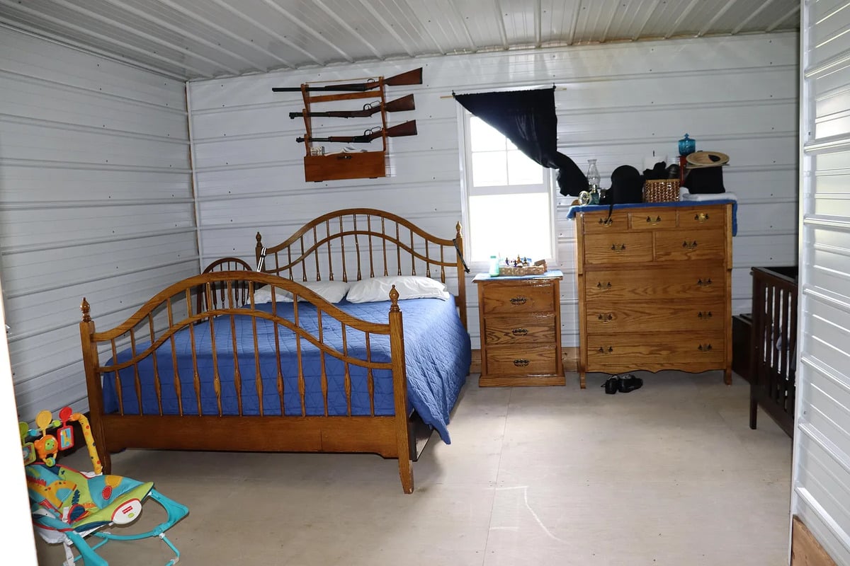 Amish bedroom with three long guns mounted on a wall rack above the bed, and a crib in the corner