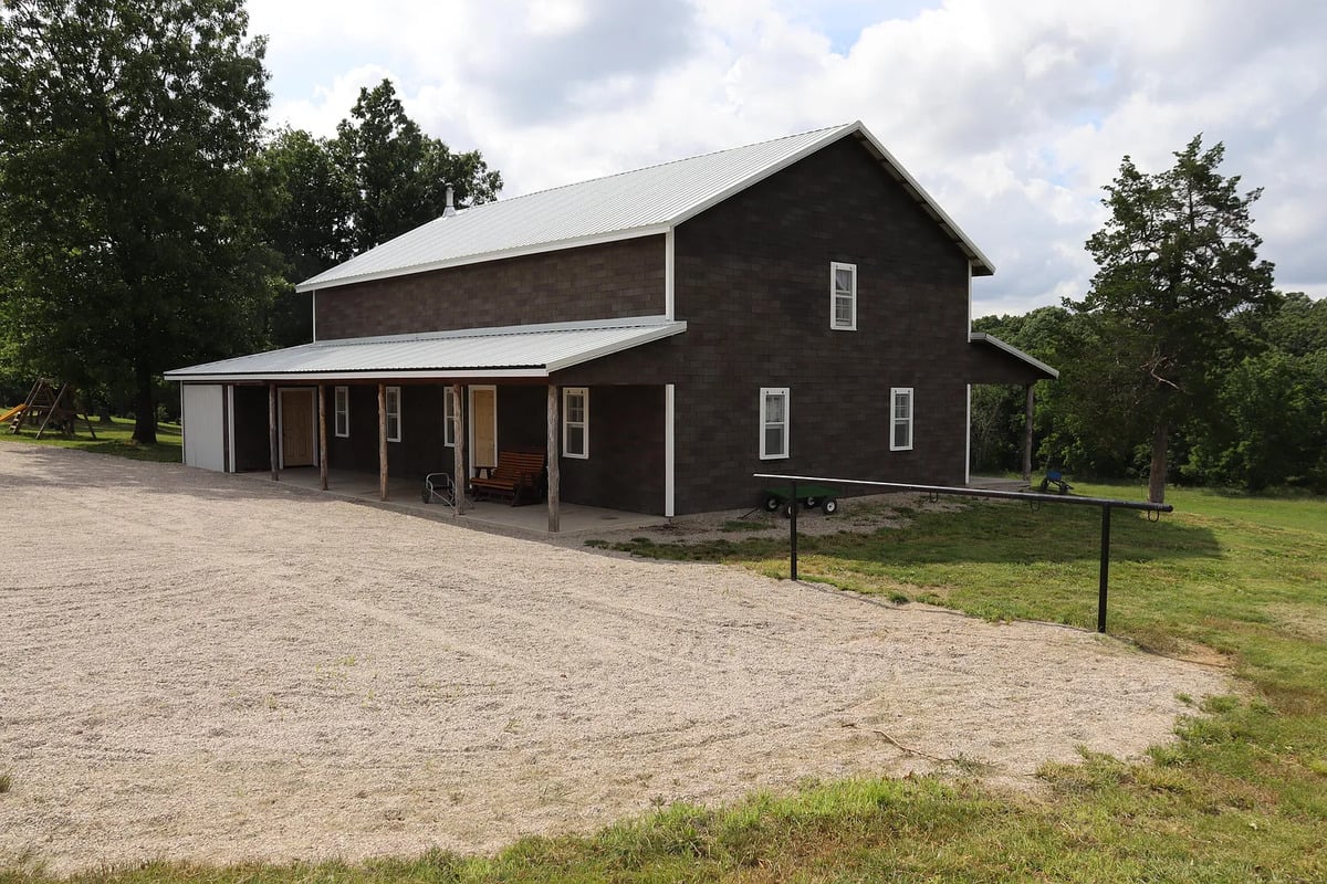 Two-story dark brown house with a metal roof and open front porch on a gravel driveway in a rural setting.