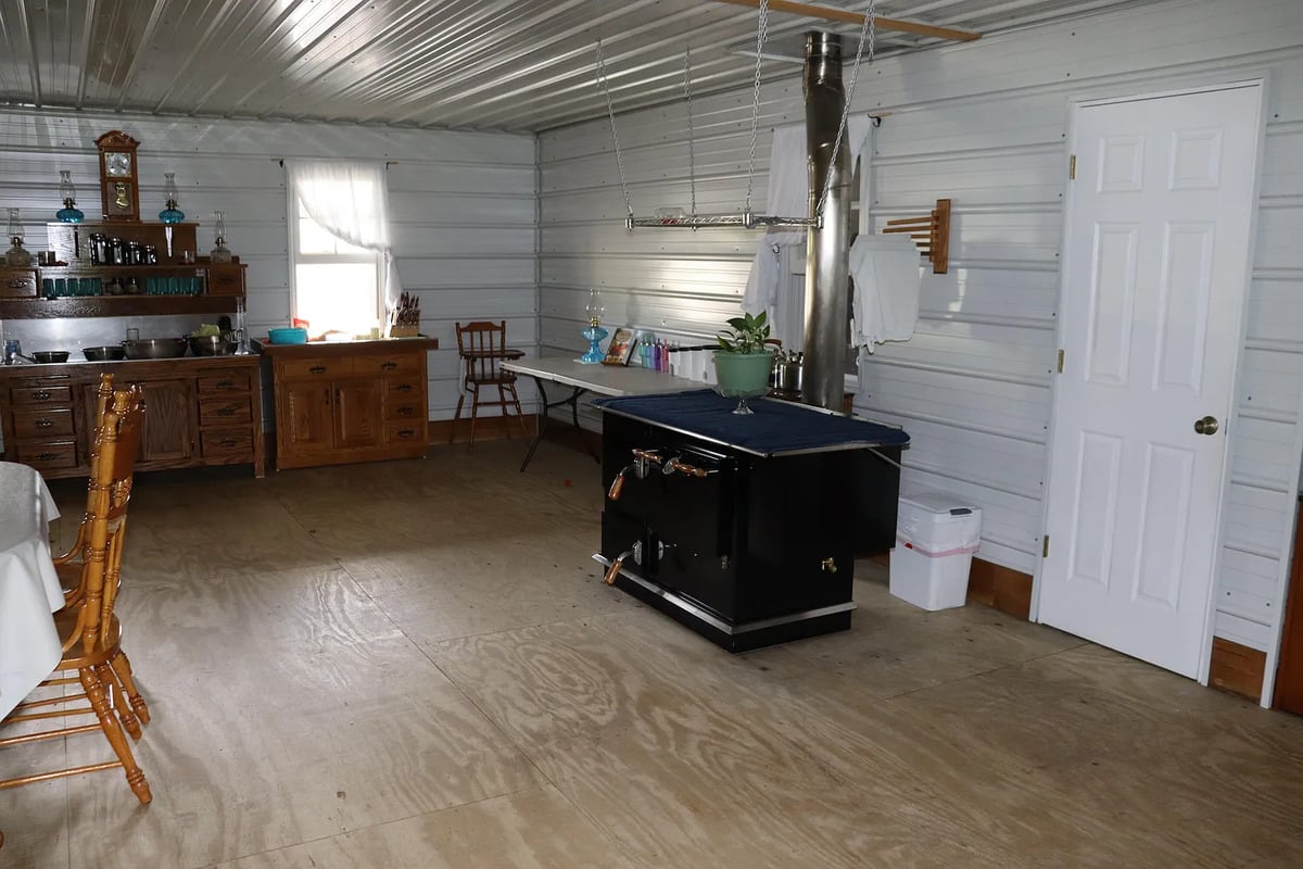 Open main room of an Amish home with a black wood cookstove, metal walls, and bare plywood floor
