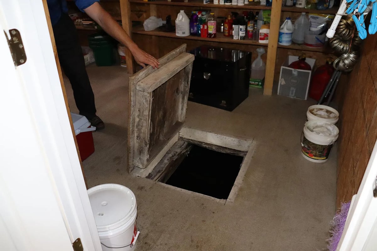 A man lifts a rough wooden hatch in the pantry floor, opening onto a dark hole below