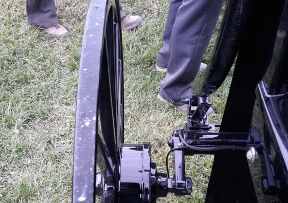 Close-up of a black trailer wheel and hitch on a grassy field with people standing nearby.
