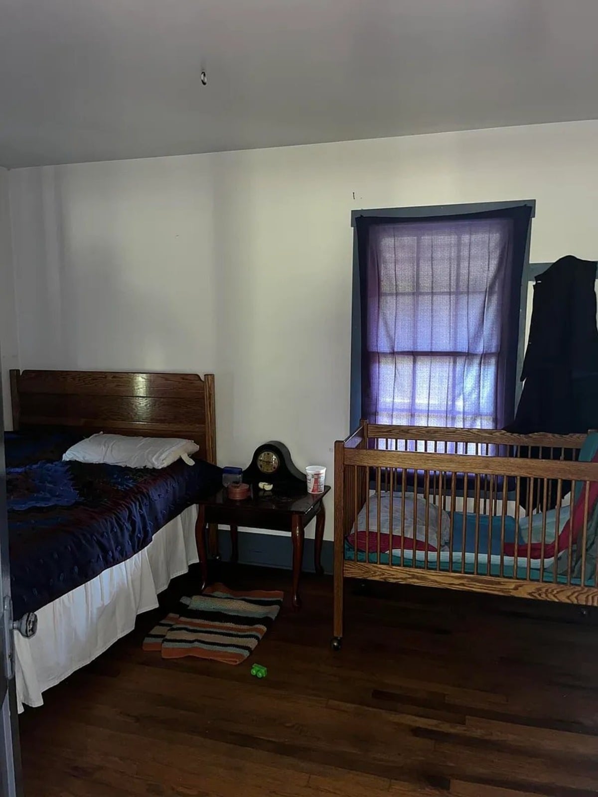 Amish bedroom with wooden bed frame, dark blue quilt, wooden crib, mantel clock, and rag rug, Randolph Mississippi