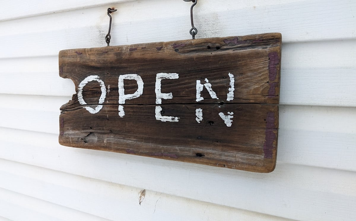 Hand-painted rustic wooden "Open" sign hanging on white siding at Peachey's Country Store, an Amish bakery in Somerset County, PA.
