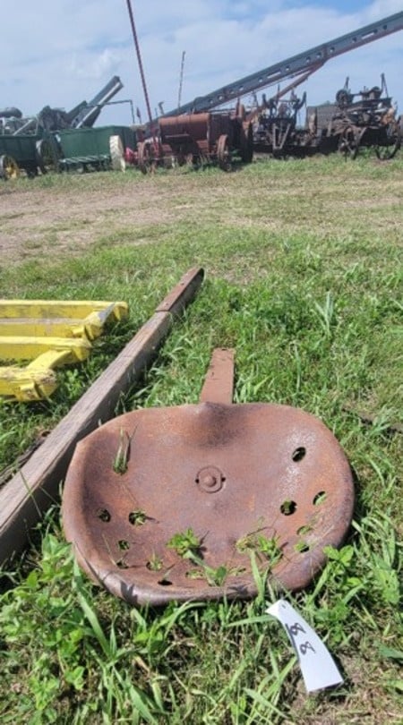 Rusted stamped-iron implement seat lying in the grass with an auction tag