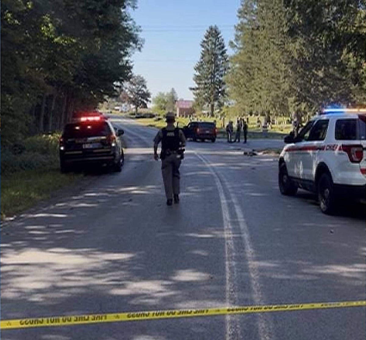 A state trooper walks down a tree-lined rural road behind yellow police tape, with patrol vehicles parked on both sides and officers gathered near a cemetery ahead