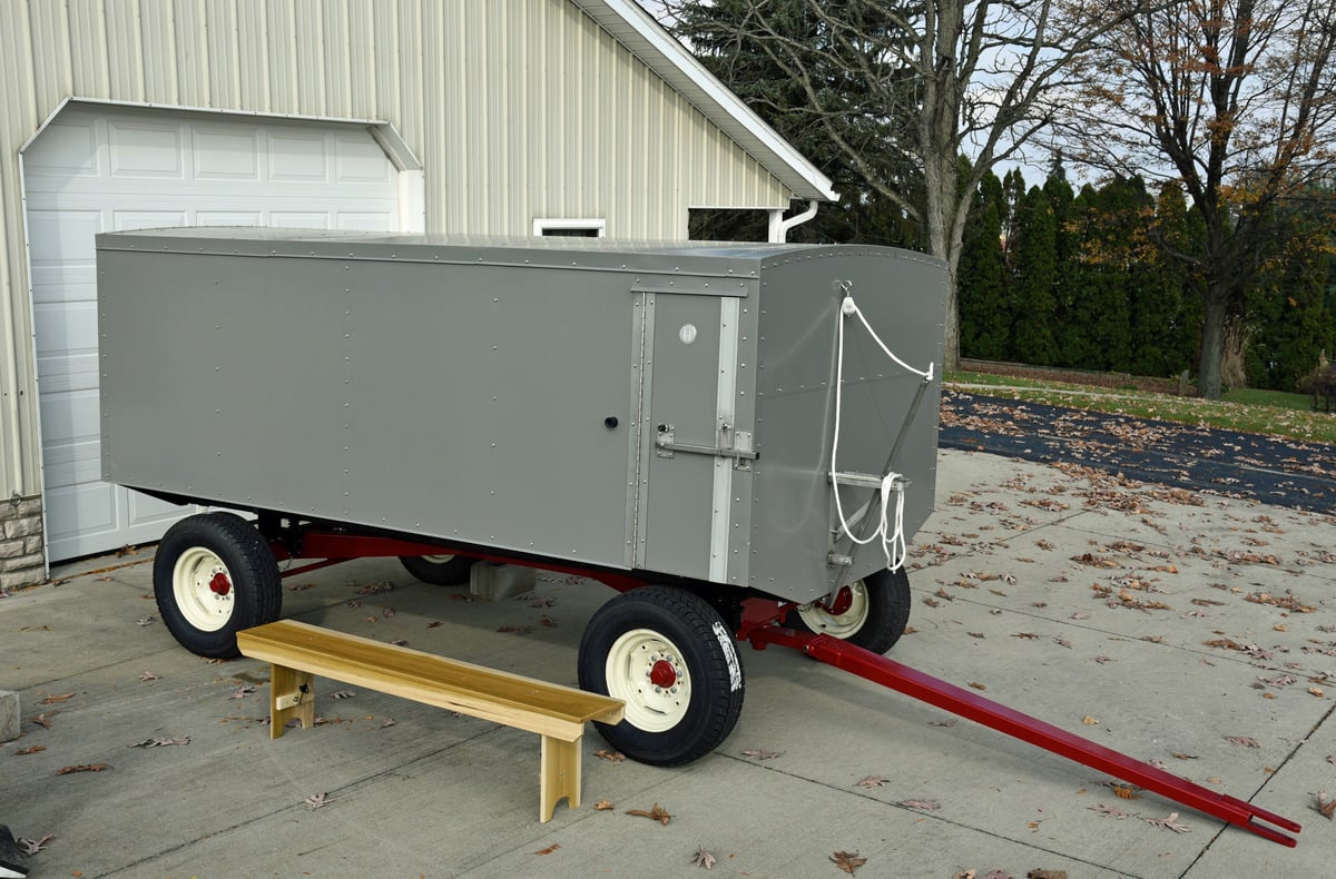 Gray enclosed bench wagon with red running gear, a wooden bench standing in front of it