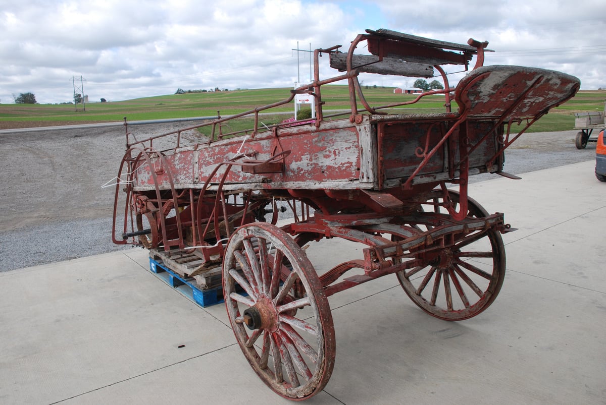 Weathered horse-drawn fire hose wagon with peeling paint and bare wood, sitting on a pallet