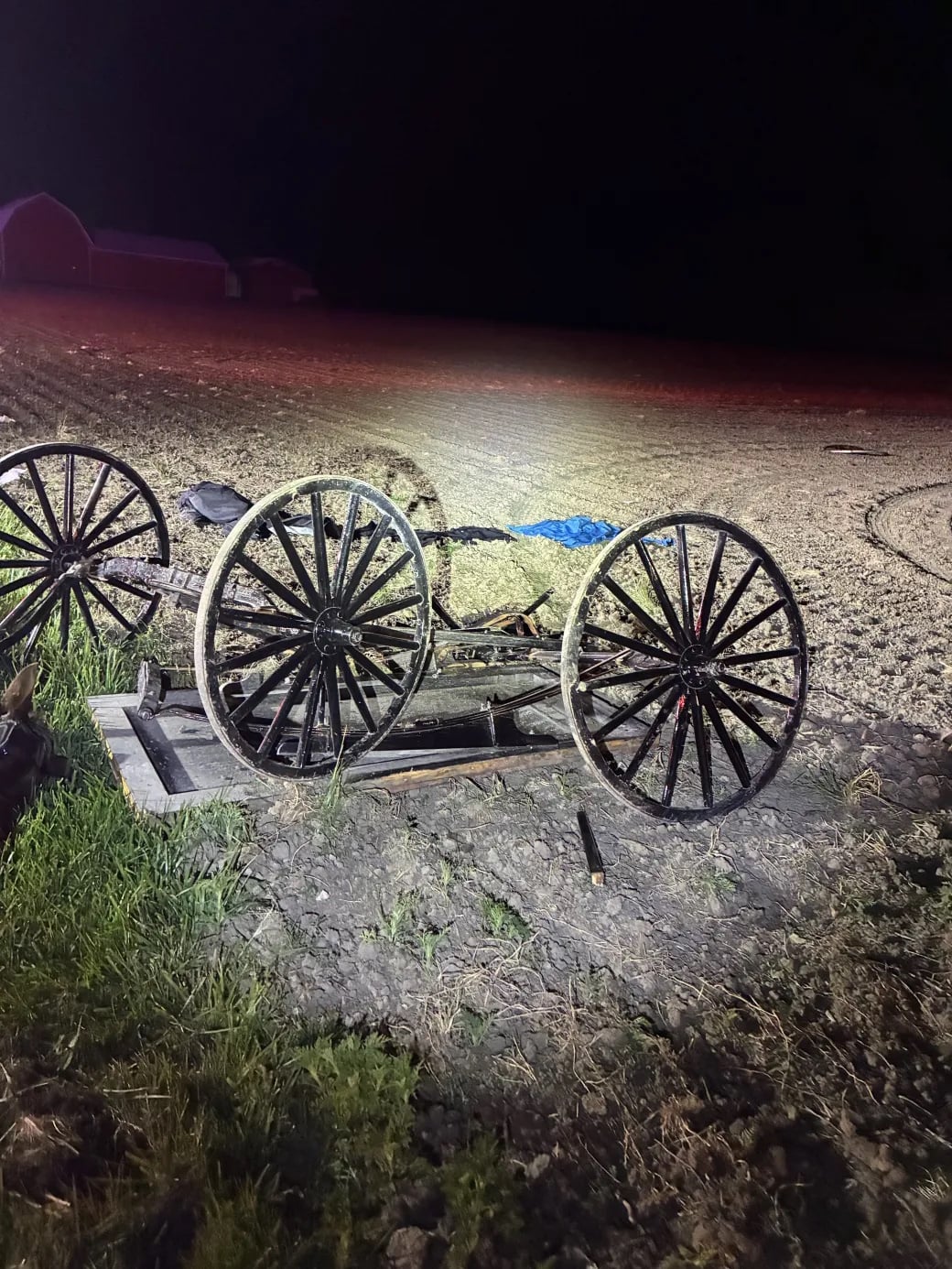Full view of Wrecked Amish Buggy Adams County Barn in Background Old wooden wagon frame with three large spoked wheels lying on a dirt and gravel area beside a field at night.