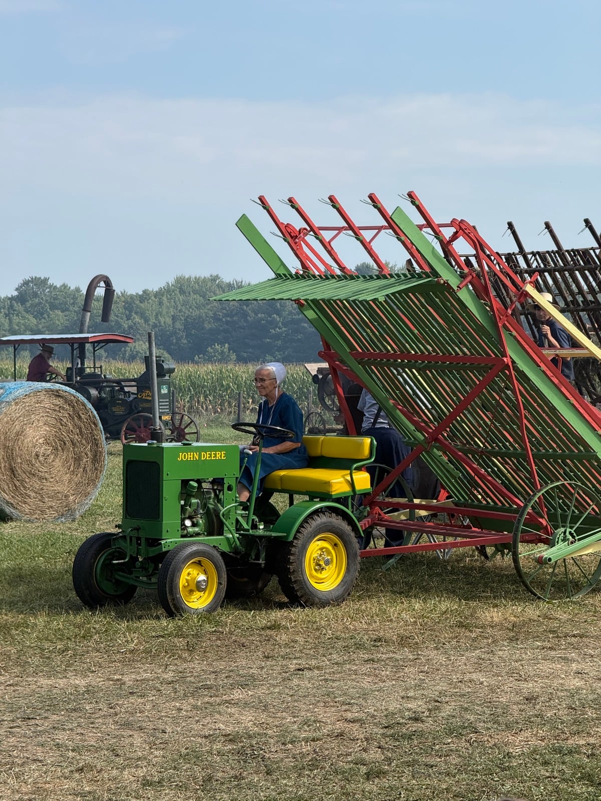 Older Amish woman drives a small John Deere tractor near a hay loader at Rentown Old Fashion Days