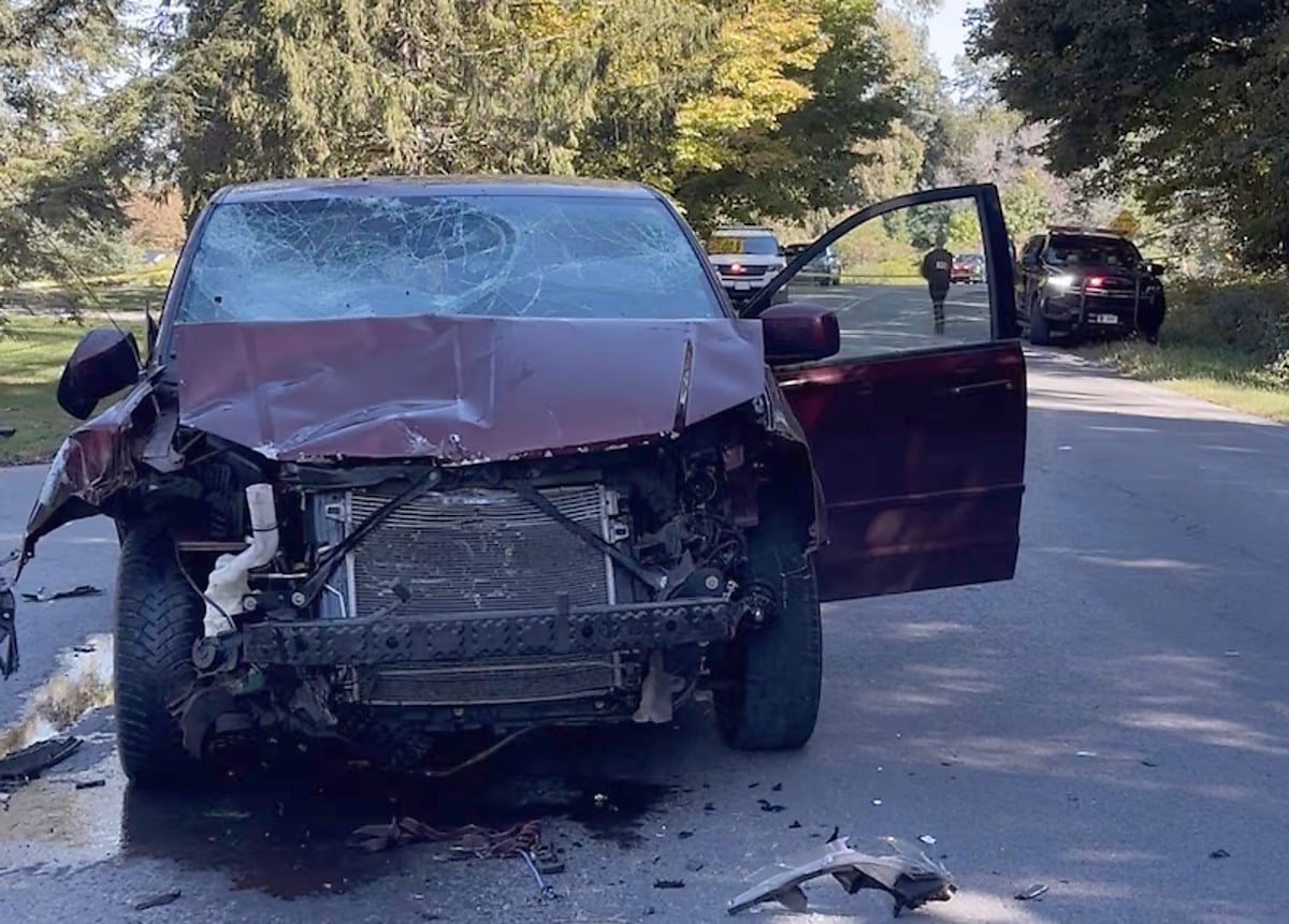 A maroon minivan with a shattered windshield, crumpled hood, and front bumper torn away sits on a rural road with its driver's door open, debris scattered on the pavement and police vehicles parked behind it
